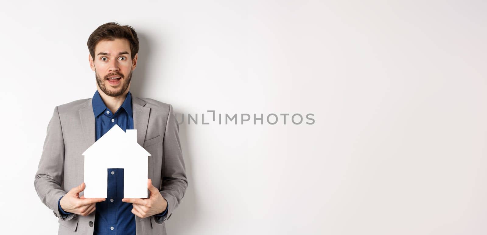 Real estate. Excited business man in suit holding paper house cutout and looking amazed at camera, buying apartment, white background.