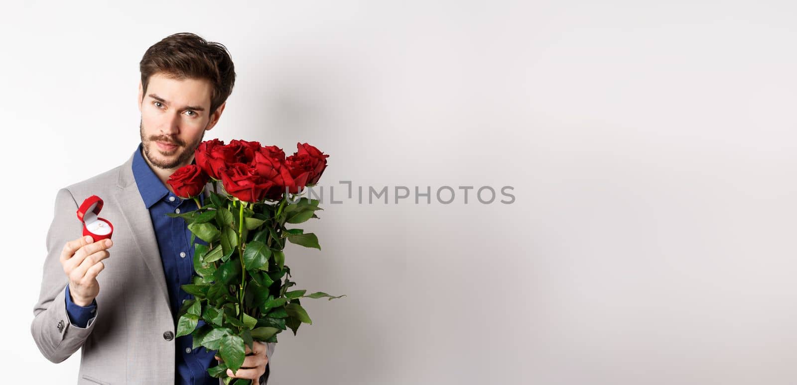 Handsome man in suit, showing engagement ring and looking romantic at camera, standing with red roses over white background. Valentines day and love concept by Benzoix