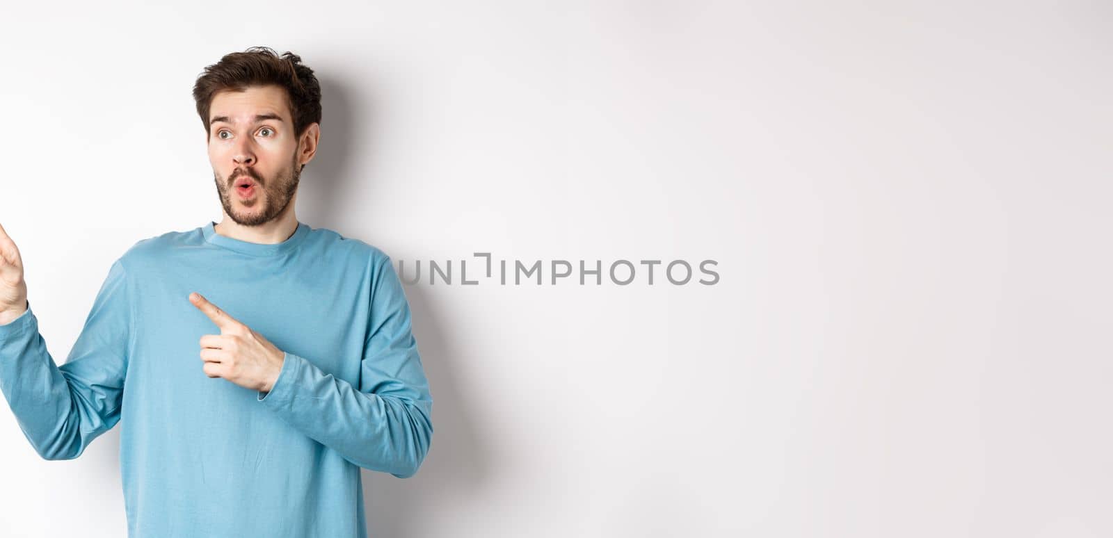 Wow check it out. Impressed young man pointing and looking left at special promo offer or discounts on white background, gasping amazed, standing in studio.