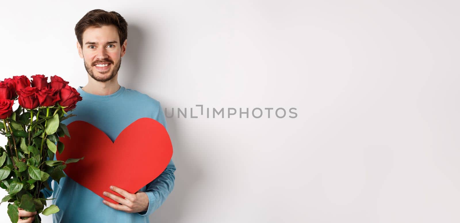 Romantic people. Handsome smiling man holding bouquet of roses and big red heart, going on Valentines day date with girlfriend, standing over white background by Benzoix