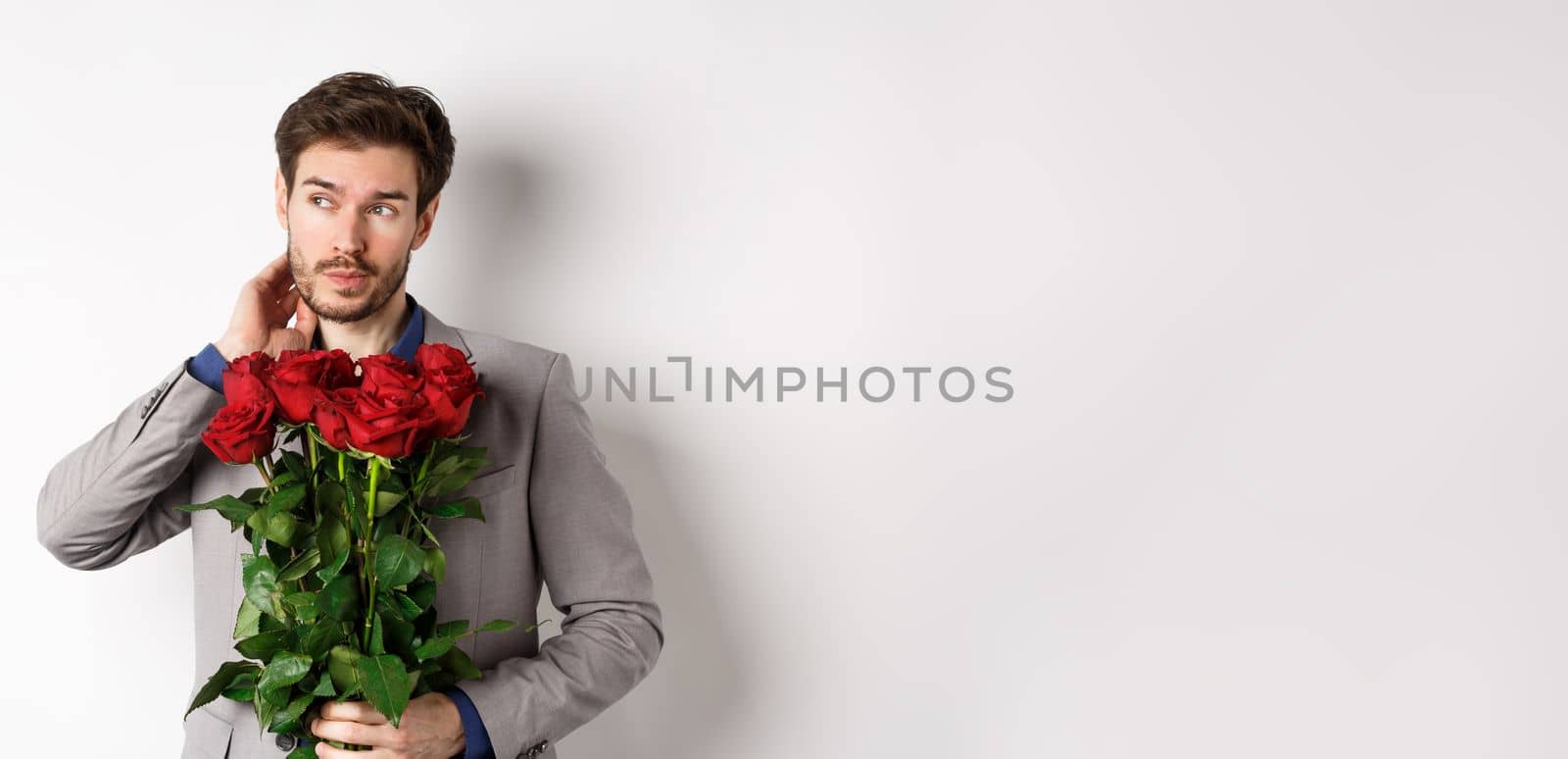 Pensive young man in suit holding bouquet of flowers, waiting for date on Valentines day, standing over white background by Benzoix