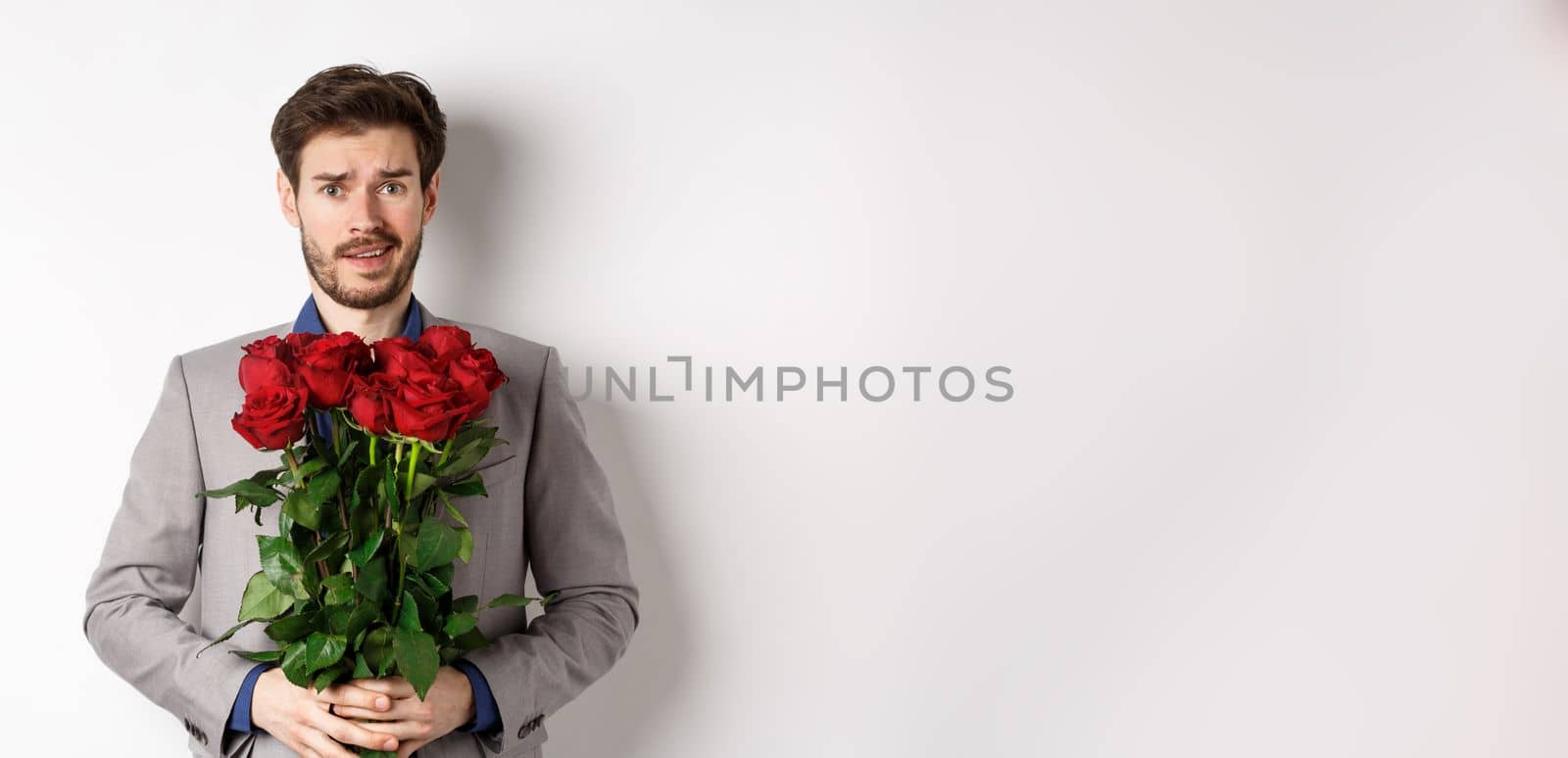 Worried boyfriend in suit, holding flowers roses and looking doubtful at camera, standing with bouquet on valentines day against white background by Benzoix