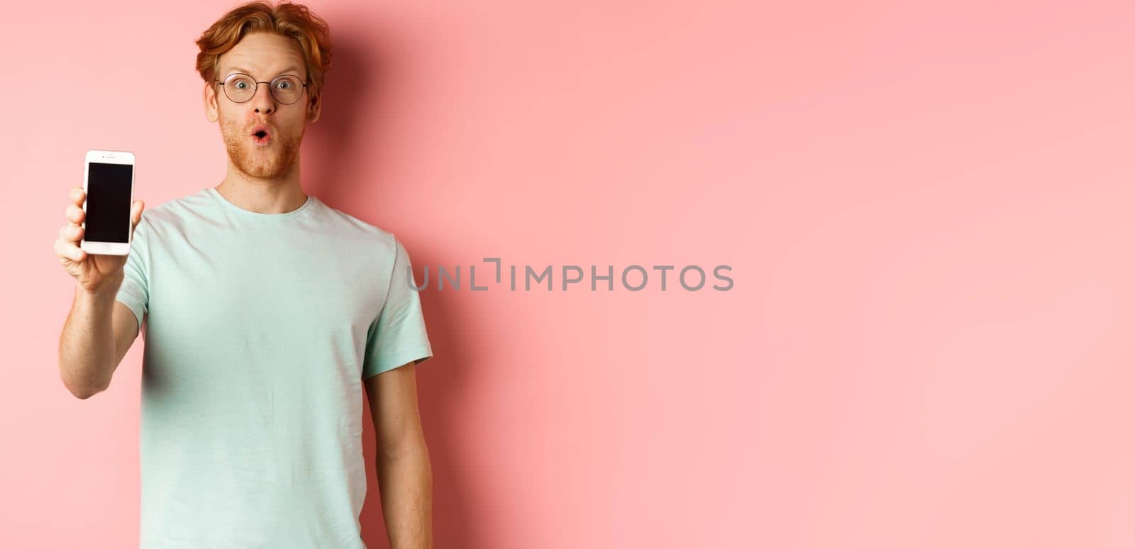 Image of handsome guy with red hair, wearing glasses and t-shirt, saying wow and showing smartphone screen, standing against pink background by Benzoix