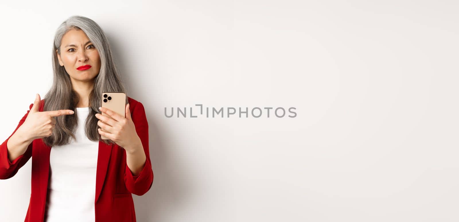 Disappointed mature asian woman complaining, pointing finger at smartphone and looking displeased, standing over white background by Benzoix