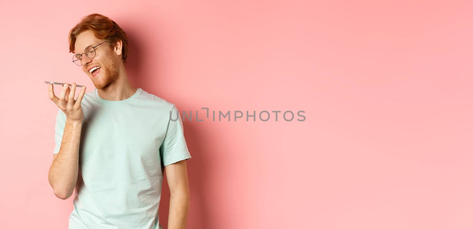 Happy young man with red hair, smiling pleased while record voice message on smartphone, talking to virtual assistant, standing over pink background by Benzoix
