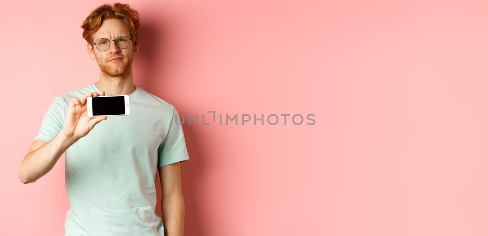 Skeptical redhead guy in glasses showing smartphone screen horizontally, smirk and frowning disappointed, standing over pink background by Benzoix