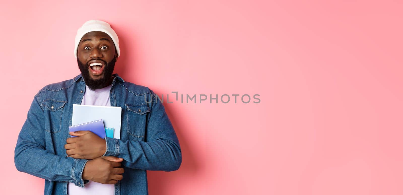 Image of adult african-american man holding notebooks and smiling, studying at courses, standing over pink background.