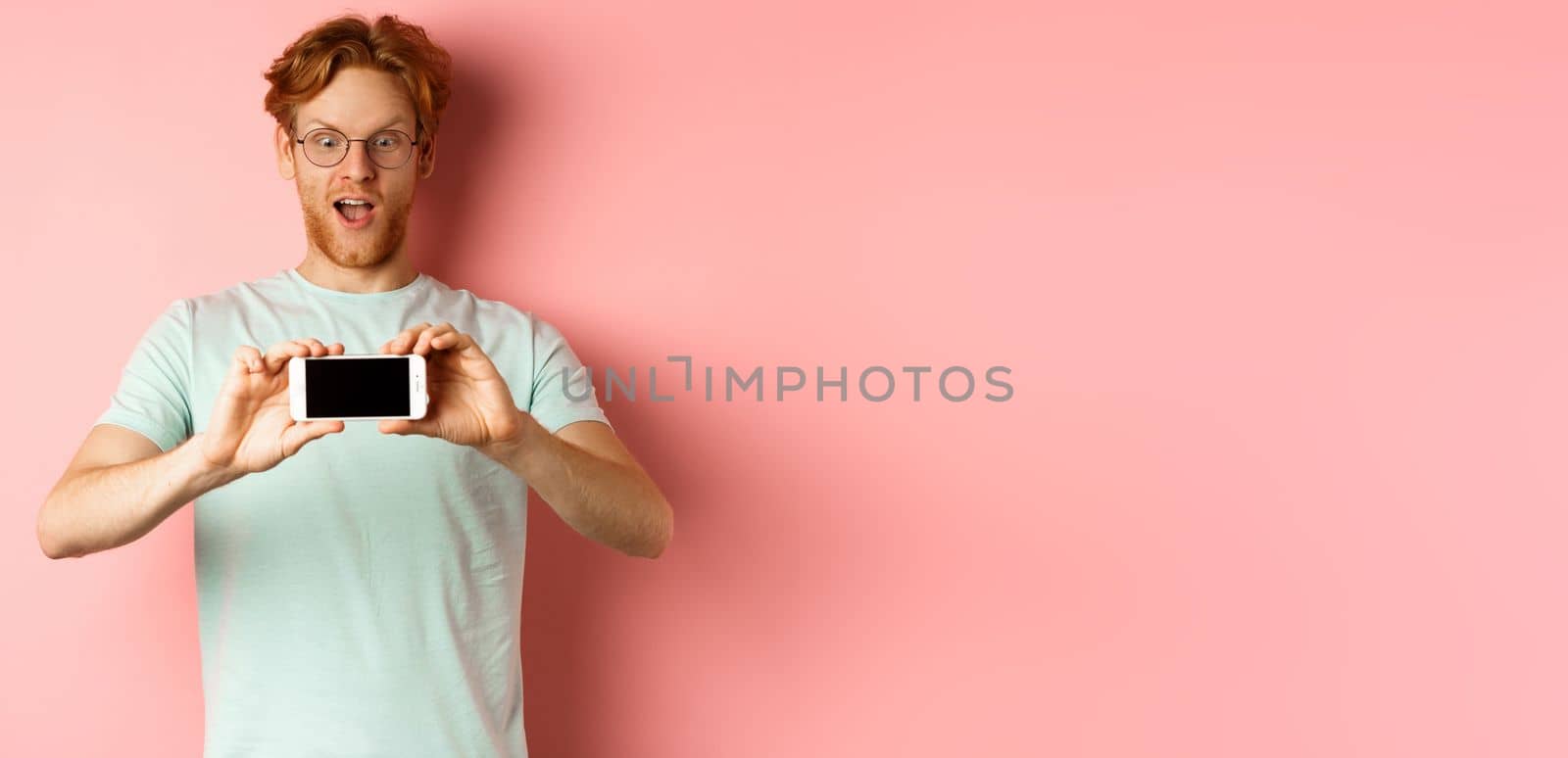 Amazed redhead man gasping and staring with awe phone, showing blank smartphone screen horizontally, standing over pink background by Benzoix
