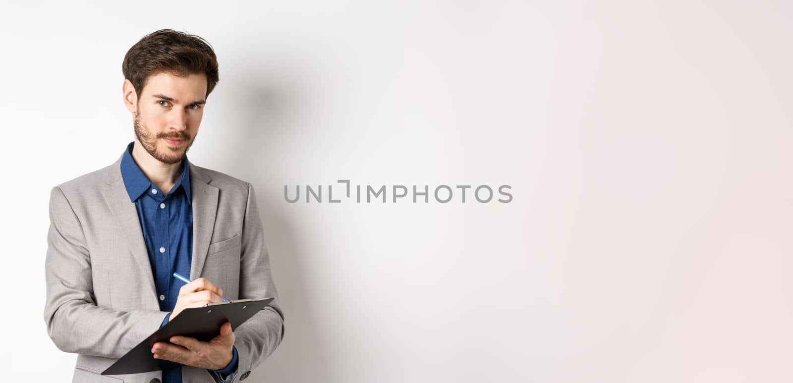 Successful businessman in grey suit taking notes, working in office clothing, holding clipboard and looking at camera, white background.