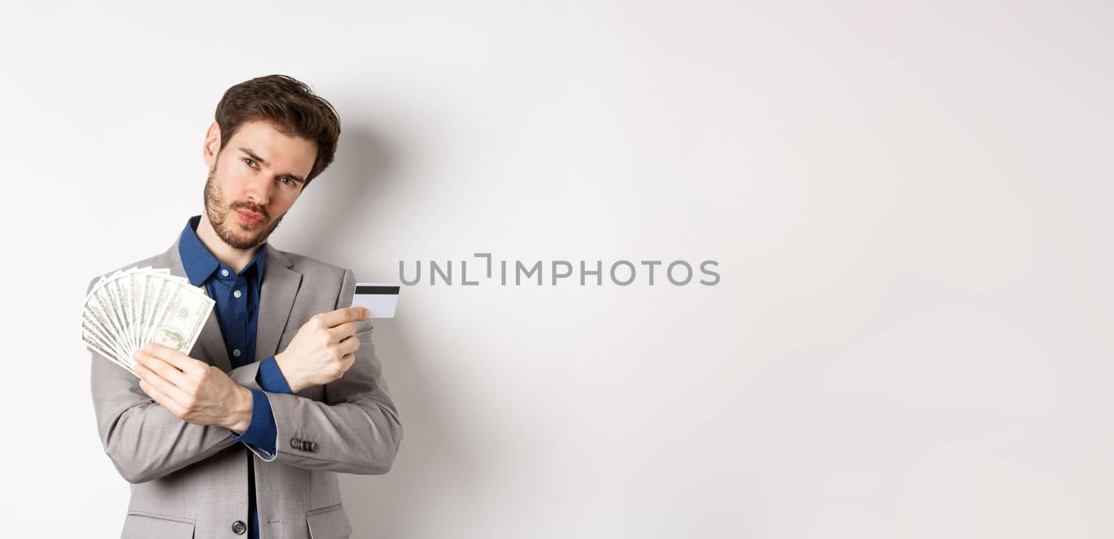 Cool young rich man in suit showing dollar bills and plastic credit card, making money, standing on white background by Benzoix