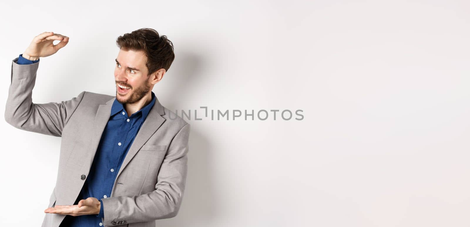 Smiling happy businessman in suit showing large size object, shaping big thing with hands and look pleased, standing on white background by Benzoix