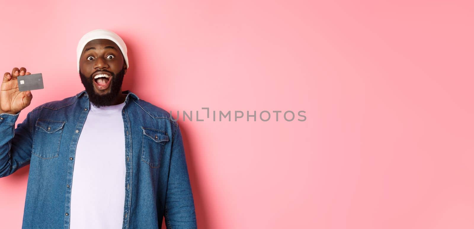 Shopping concept. Amazed Black guy showing credit card, staring at camera impressed, recommending bank, standing over pink background.