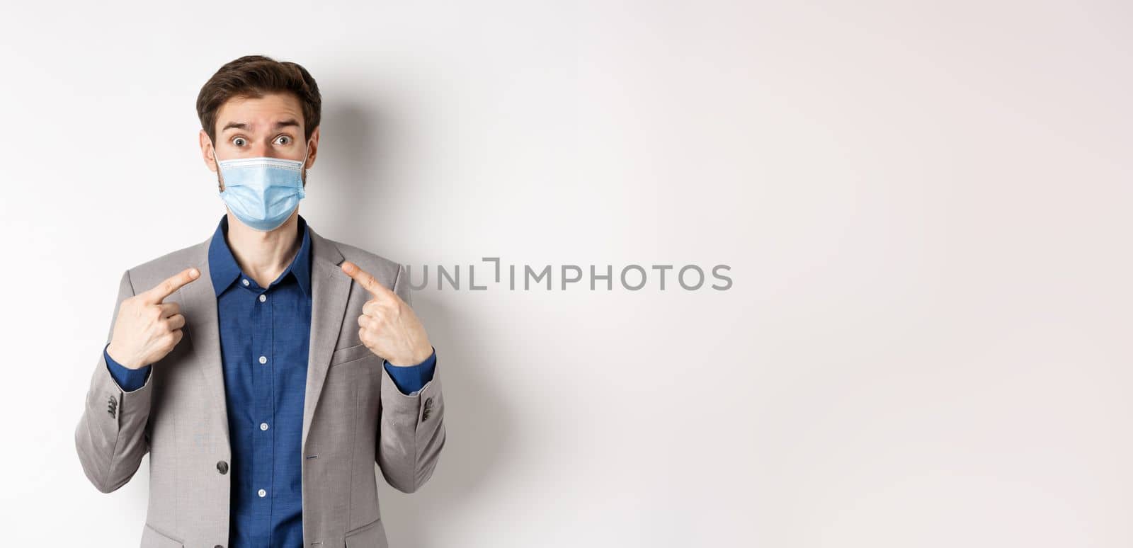 Covid-19, pandemic and business concept. Excited businessman in suit pointing at his medical mask and looking at camera, white background by Benzoix