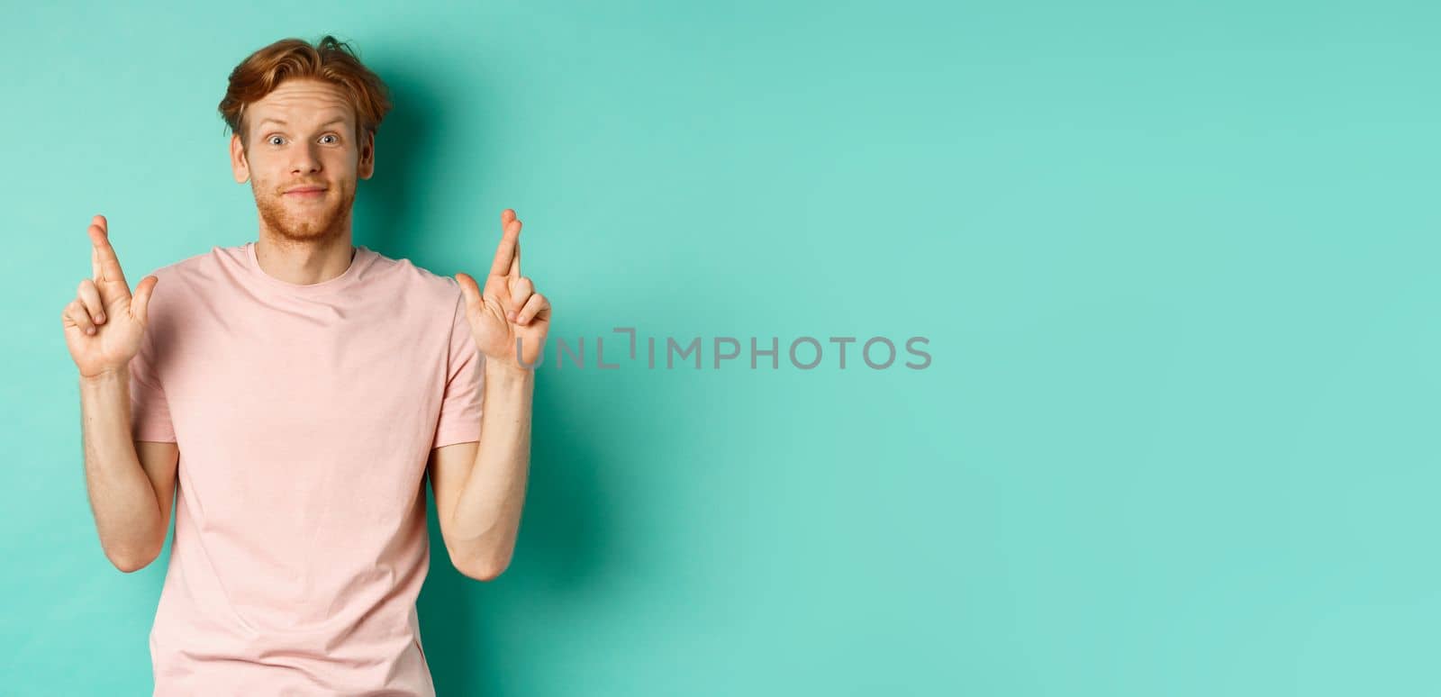 Smiling hopeful man with red hair making a wish, cross fingers for good luck and expecting something good, standing over turquoise background.