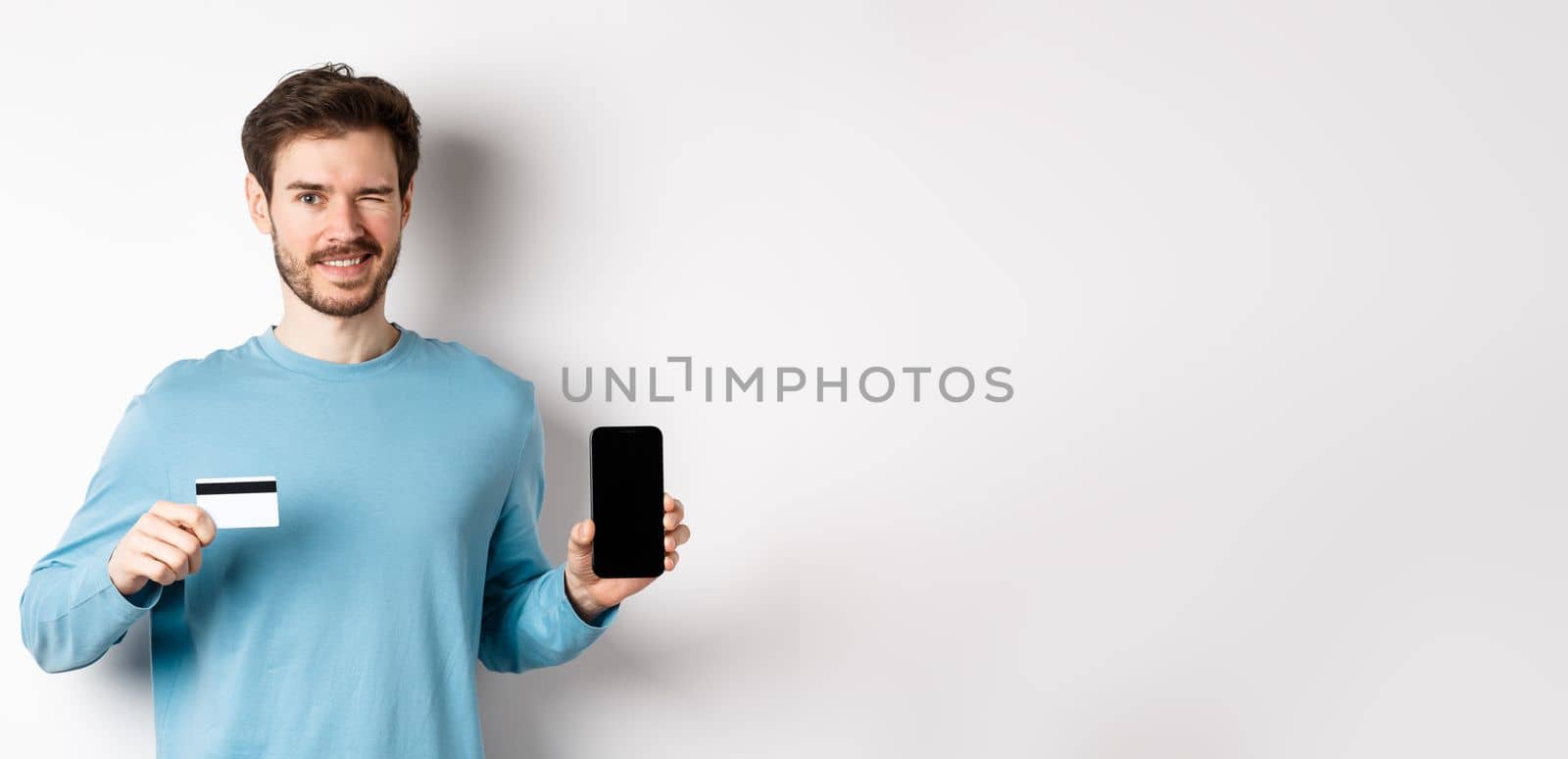 Young man in casual shirt showing empty smartphone screen and plastic credit card, winking and smiling at camera, standing on white background.