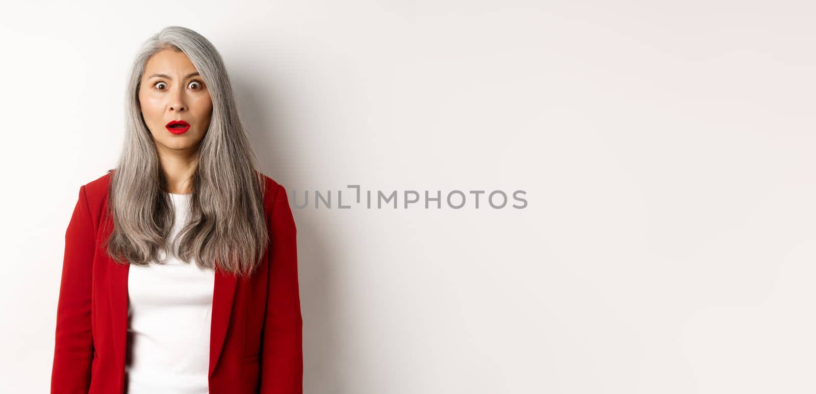 Business concept. Shocked senior female employer staring at camera speechless, standing in red blazer and makeup over white background by Benzoix