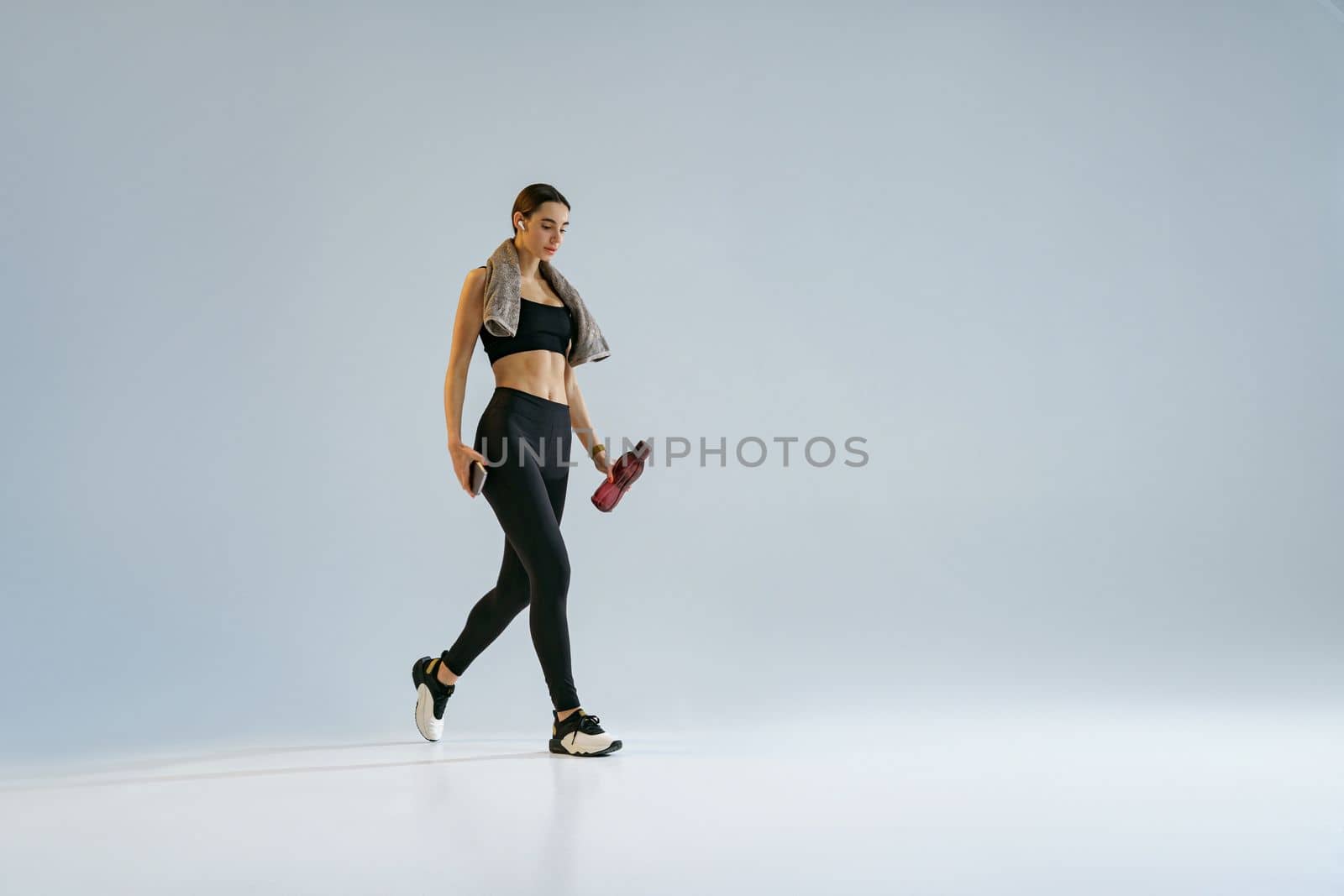 Woman walking with water bottle and towel around her neck over studio background  by Yaroslav_astakhov