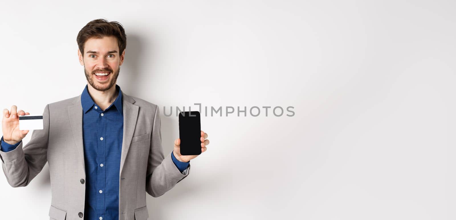 Online shopping. Smiling business man in suit showing plastic credit card with empty smartphone screen, standing against white background.