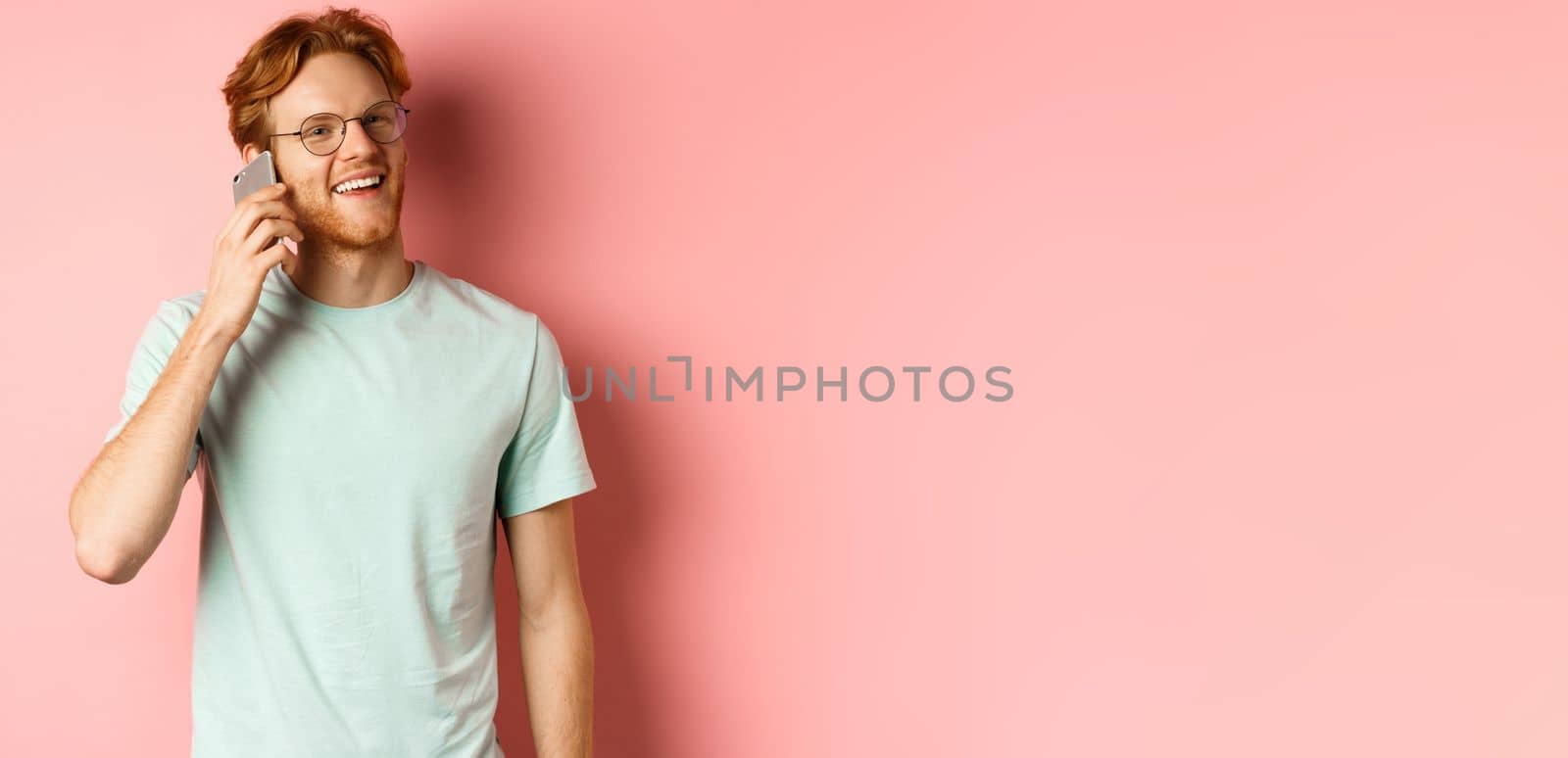 Handsomy hipster guy with red hair and beard talking on mobile phone, calling someone and looking happy, standing over pink background by Benzoix