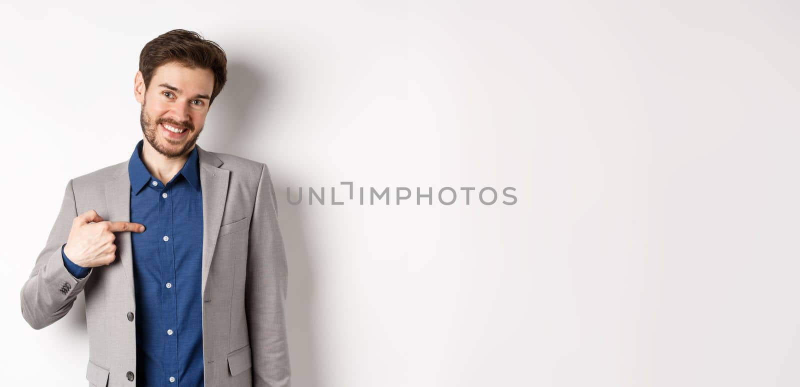 Modest smiling man in suit pointing at himself with cute face, self-promoting, standing on white background.