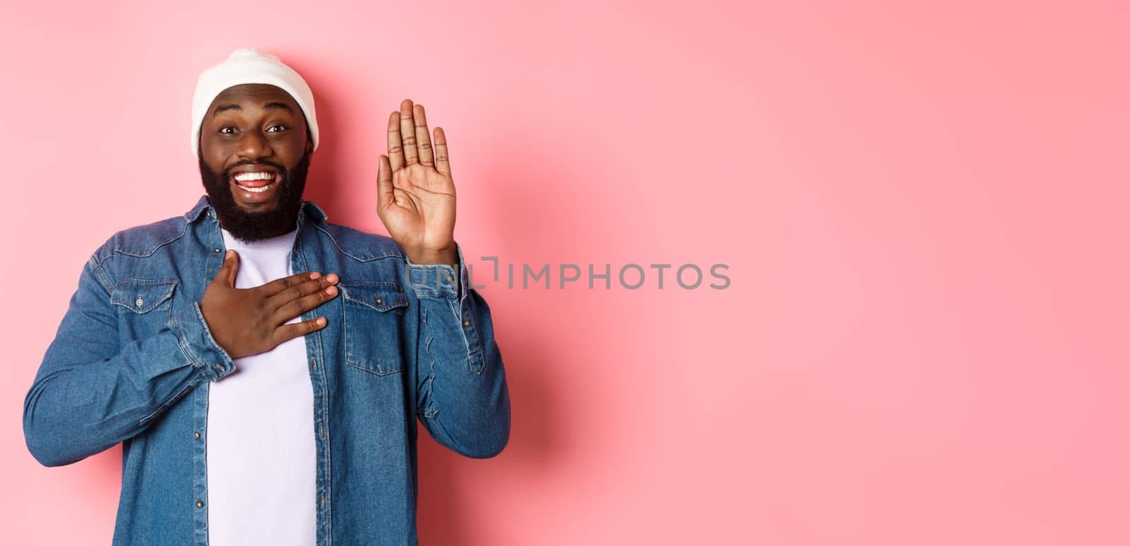 Image of smiling african-american man being honest, telling truth, hands on heart and in air, making promise or swearing, standing ove rpink background.