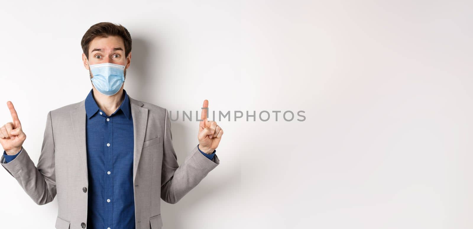 Covid-19, pandemic and business concept. Excited businessman in sterile medical mask and suit pointing fingers up, white background by Benzoix