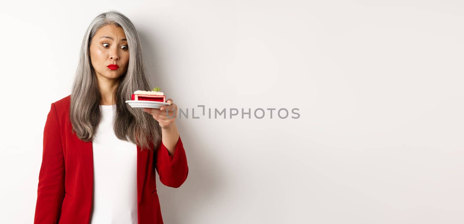 Elegant senior woman in red blazer wants take bite of sweet cake, looking with tempted face at dessert, standing over white background.