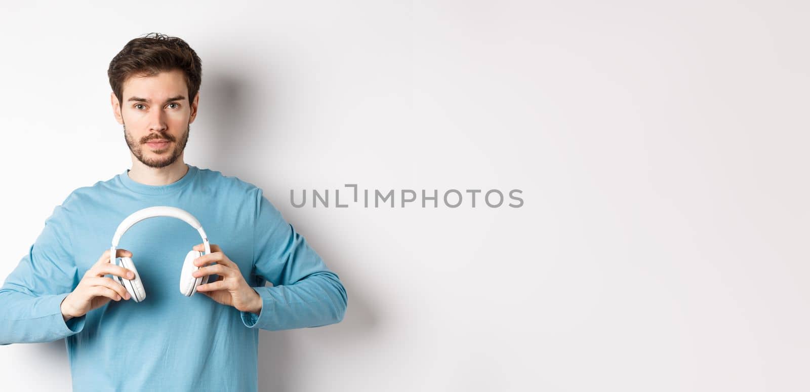 Young bearded guy in blue sweatshirt put on wireless headphones, listening music, standing on white background by Benzoix