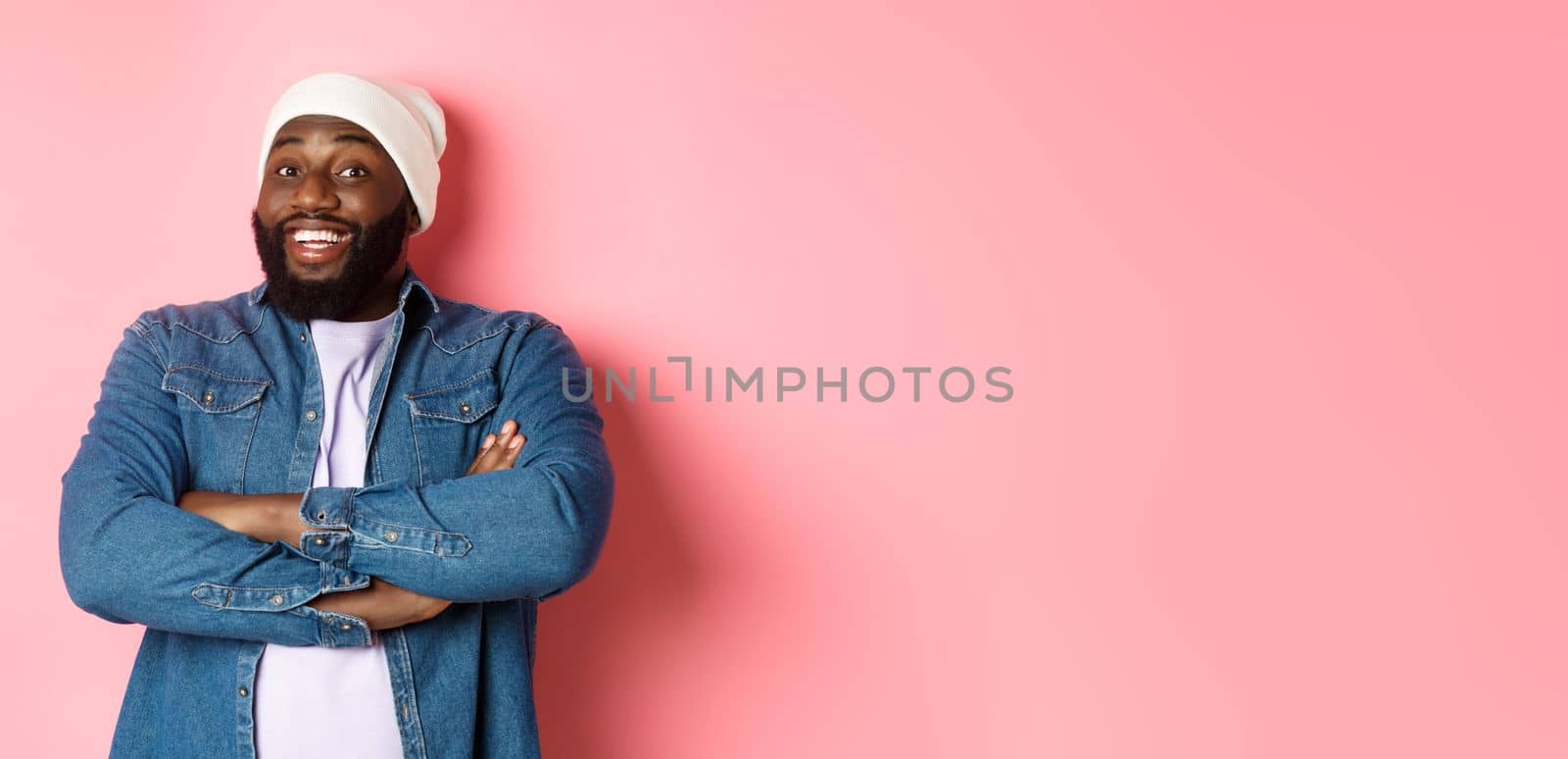 Happy Black man with beard, wearing beanie and denim shirt, looking intrigued and amused at camera, smiling with arms crossed on chest, pink background.