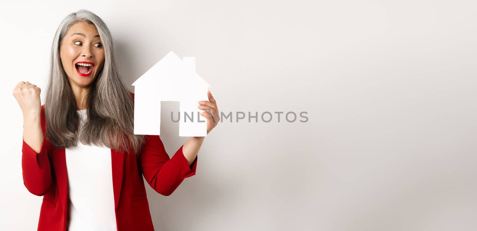 Cheerful asian senior woman buying house, scream of joy and making fist pump while showing paper house cutout, standing over white background by Benzoix