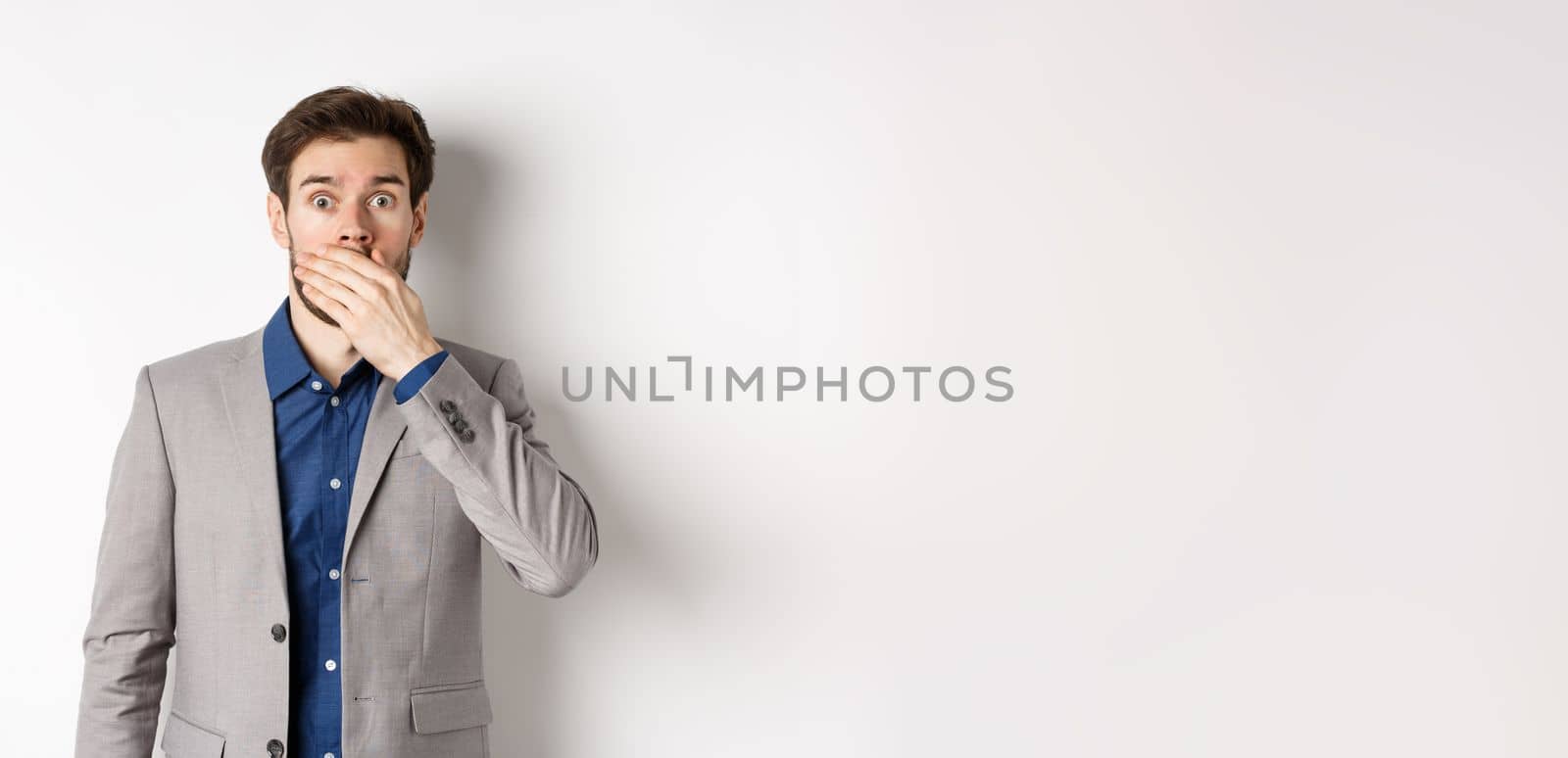 Shocked boyfriend in suit gasping, covering mouth with hand and pop eyes at camera startled, standing on white background by Benzoix