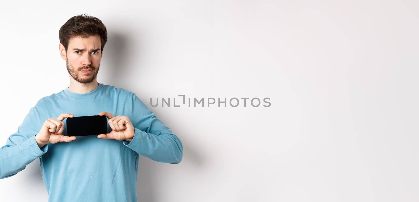 E-commerce and shopping concept. Sad and disappointed guy complaining, showing empty smartphone screen, standing on white background.