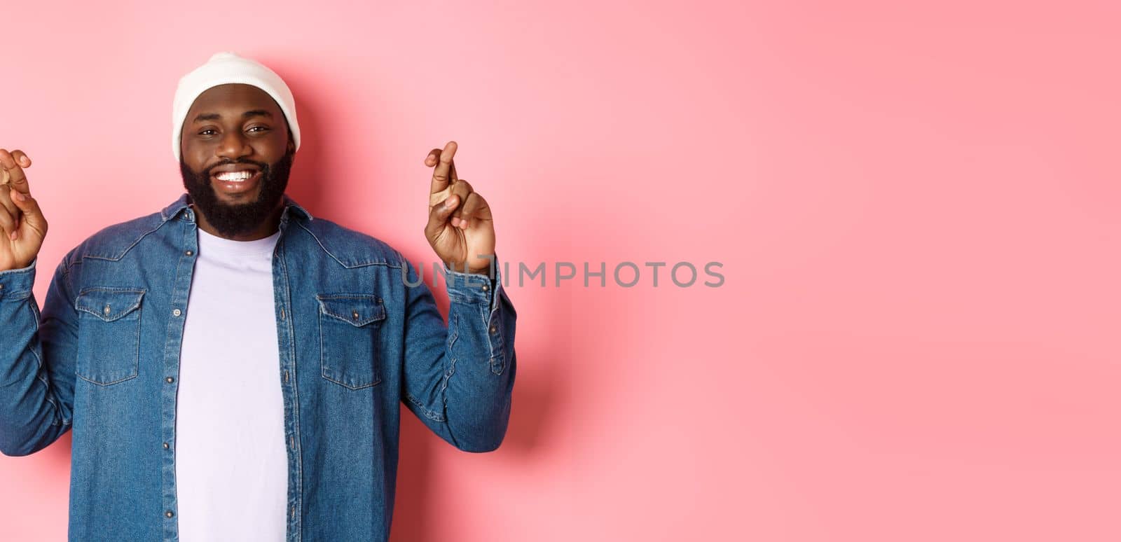 Optimistic african-american man making wish, holding fingers crossed and smiling, standing over pink background.