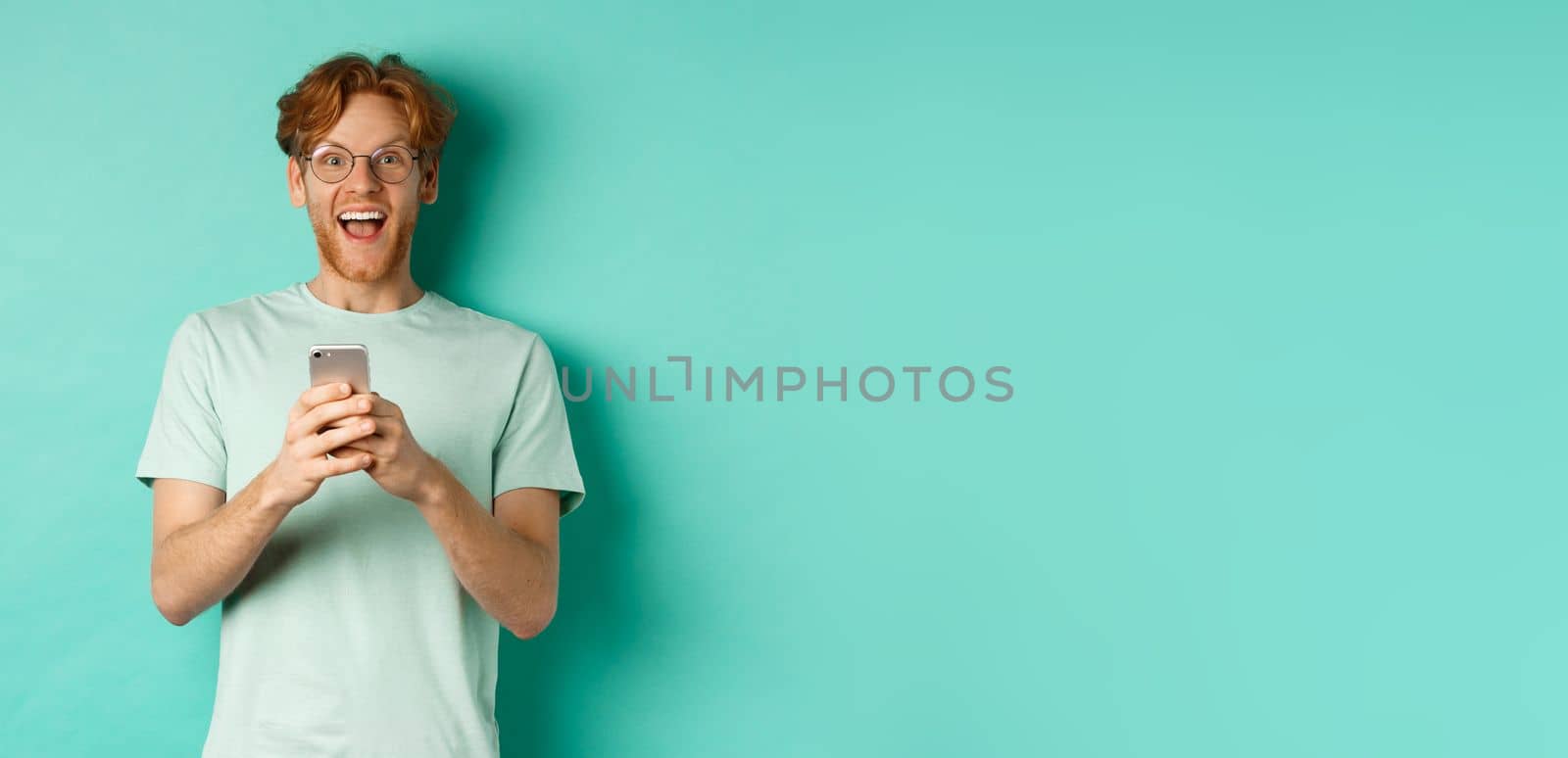 Surprised redhead man in glasses looking amazed at camera after reading promo offer on smartphone, standing against turquoise background by Benzoix