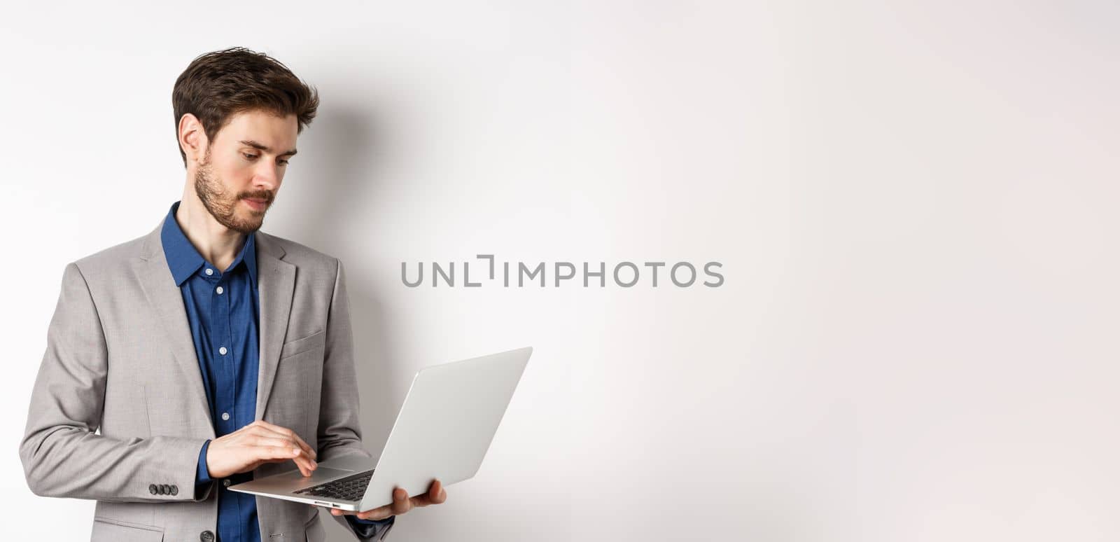 Handsome male entrepreneur working on laptop, looking serious at screen, standing against white background.
