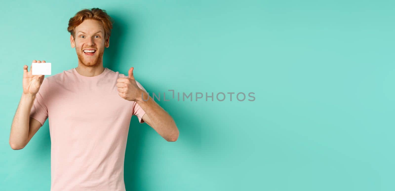 Cheerful male bank client in t-shirt showing thumb up and plastic credit card, smiling satisfied at camera, standing over turquoise background by Benzoix