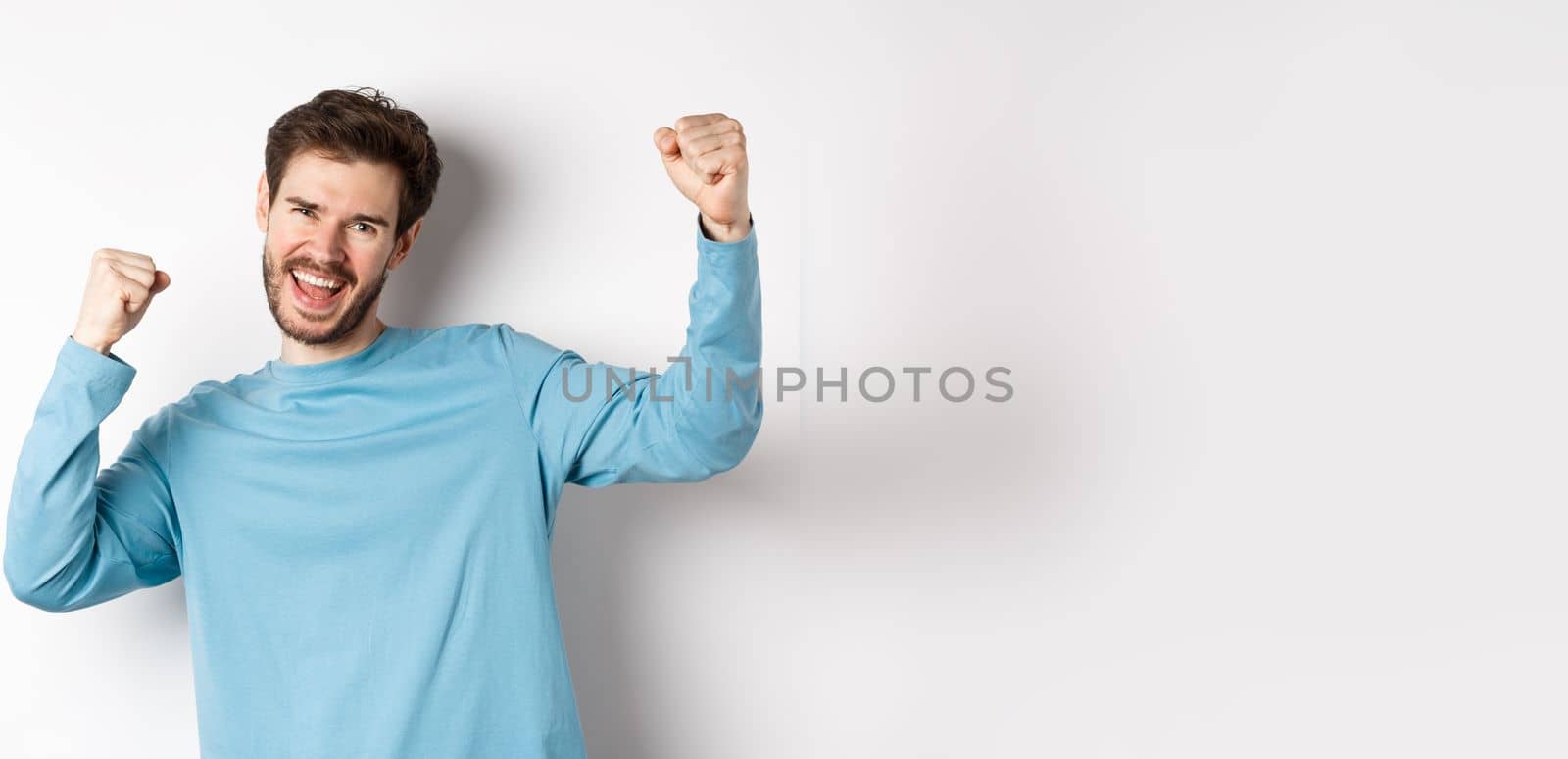 Image of happy young man triumphing, feeling like champion, winning and screaming in cheer, standing over white background.