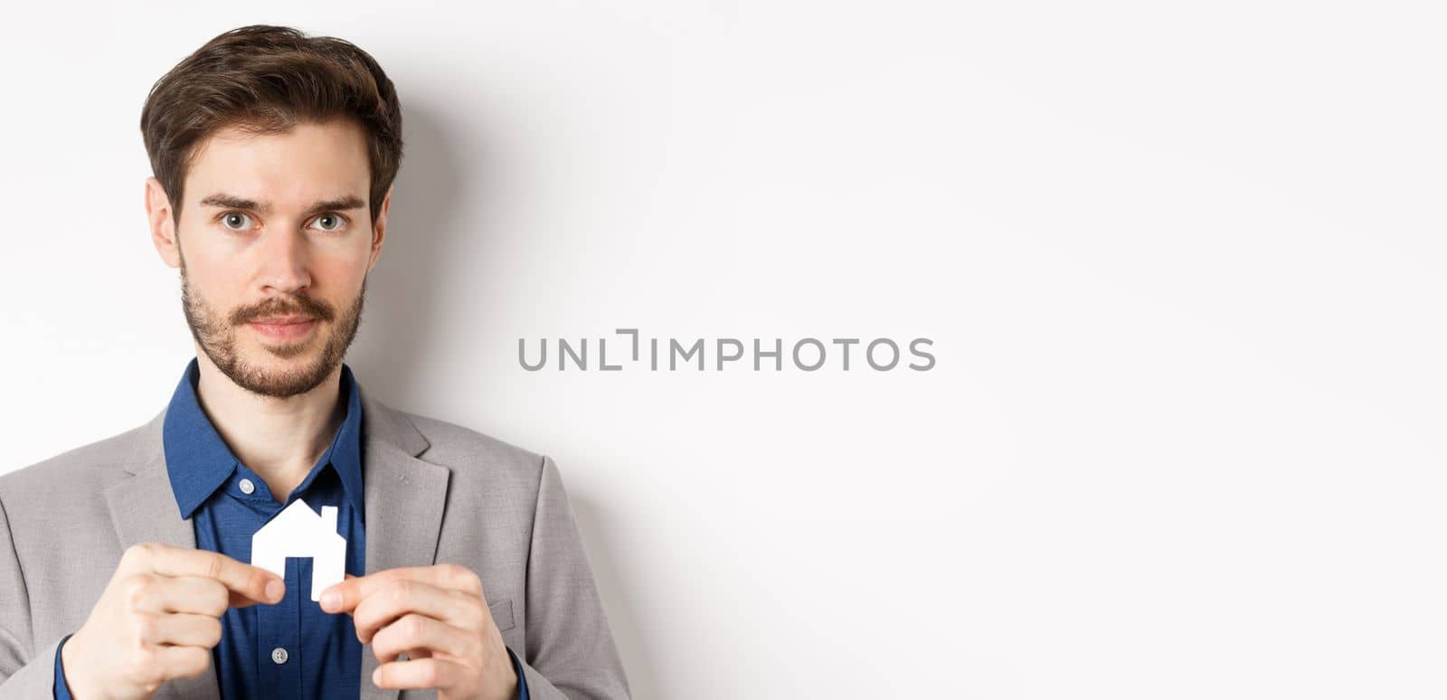 Real estate and insurance concept. Salesman realtor showing small paper house cutout and looking at camera, selling property, white background by Benzoix