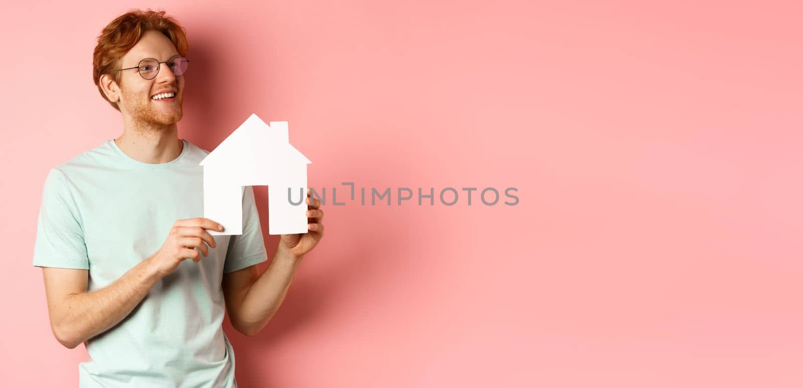 Real estate. Happy redhead man dreaming of buying property, looking right and showing paper house cutout, standing over pink background.