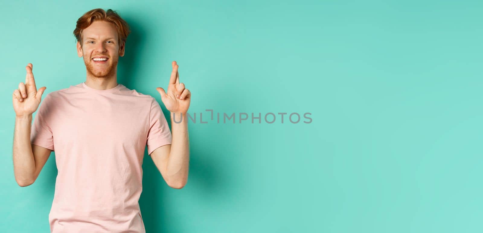 Optimistic guy with red hair and beard smiling, cross fingers for good luck and looking hopeful at camera, making a wish, standing over mint background by Benzoix