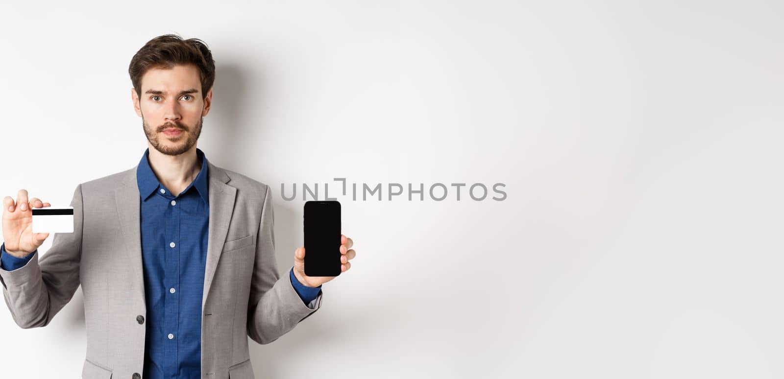 Online shopping. Serious bearded man in business suit showing plastic credit card with empty smartphone screen, standing against white background.