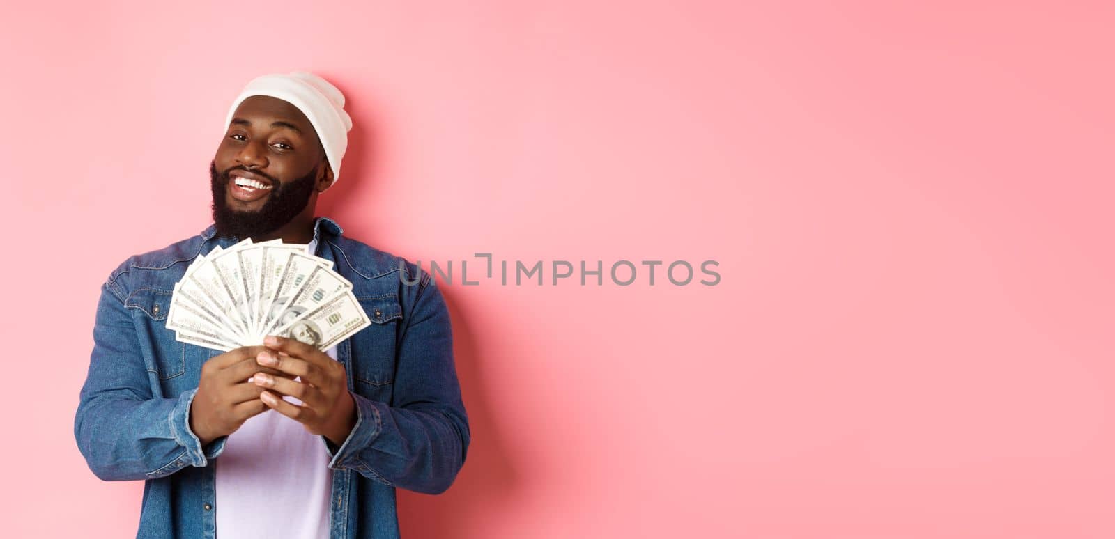 Satisfied rich african-american man showing money, smiling pleased and show-off with income, standing with dollars over pink background.