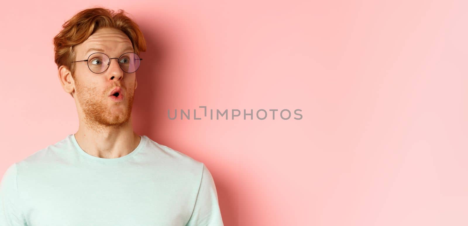 Face of intrigued young man with ginger hair and beard, raising eyebrows surpirsed and looking at upper right corner logo, standing over pink background.