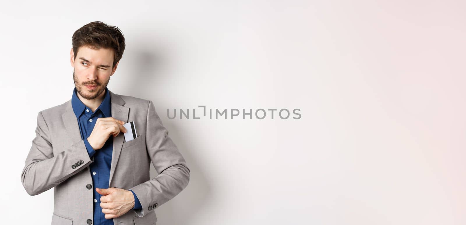 Confident and successful businessman winking aside while place credit card in suit pocket, standing on white background by Benzoix