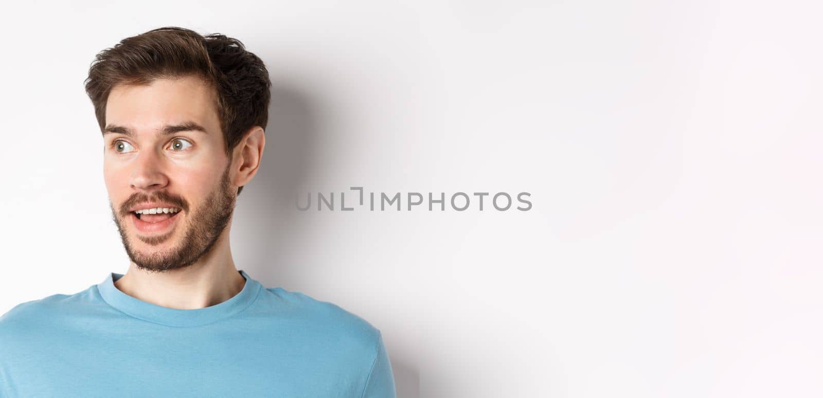 Close-up of surprised and excited bearded man, looking left with opened mouth and amazed face, checking out promo, white background.