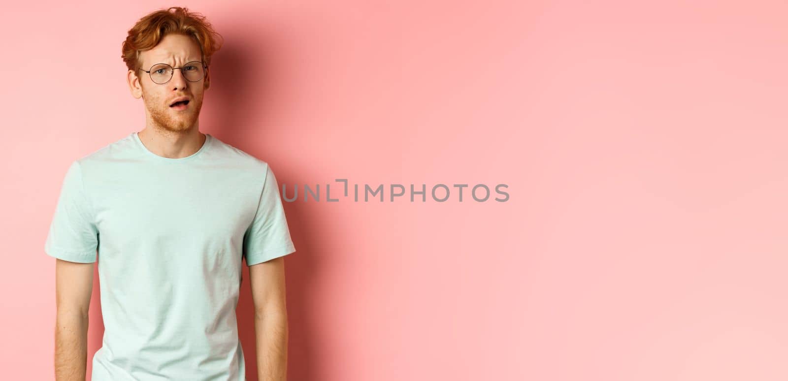 Portrait of handsome redhead man in glasses looking confused, open mouth and stare at camera, cant understand nothing, standing over pink background by Benzoix