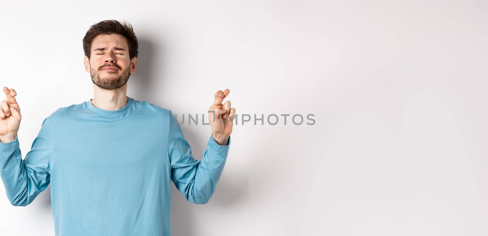Worried hopeful man praying, cross fingers for good luck with eyes closed, making wish, pleading over white background.