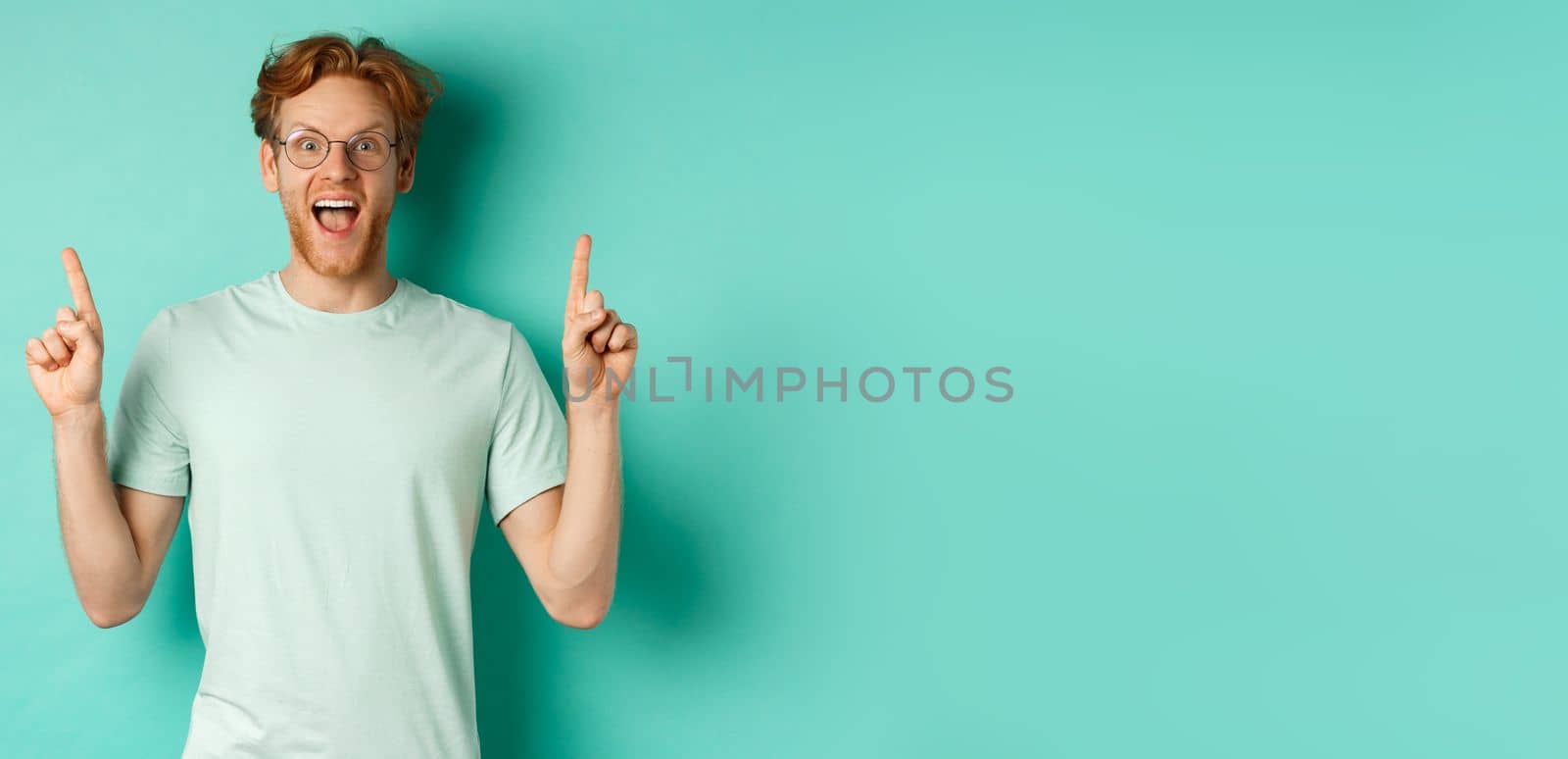 Surprised young man with ginger hair, wearing glasses and t-shirt, gasping in awe and pointing fingers up at promo deal, standing over mint background by Benzoix
