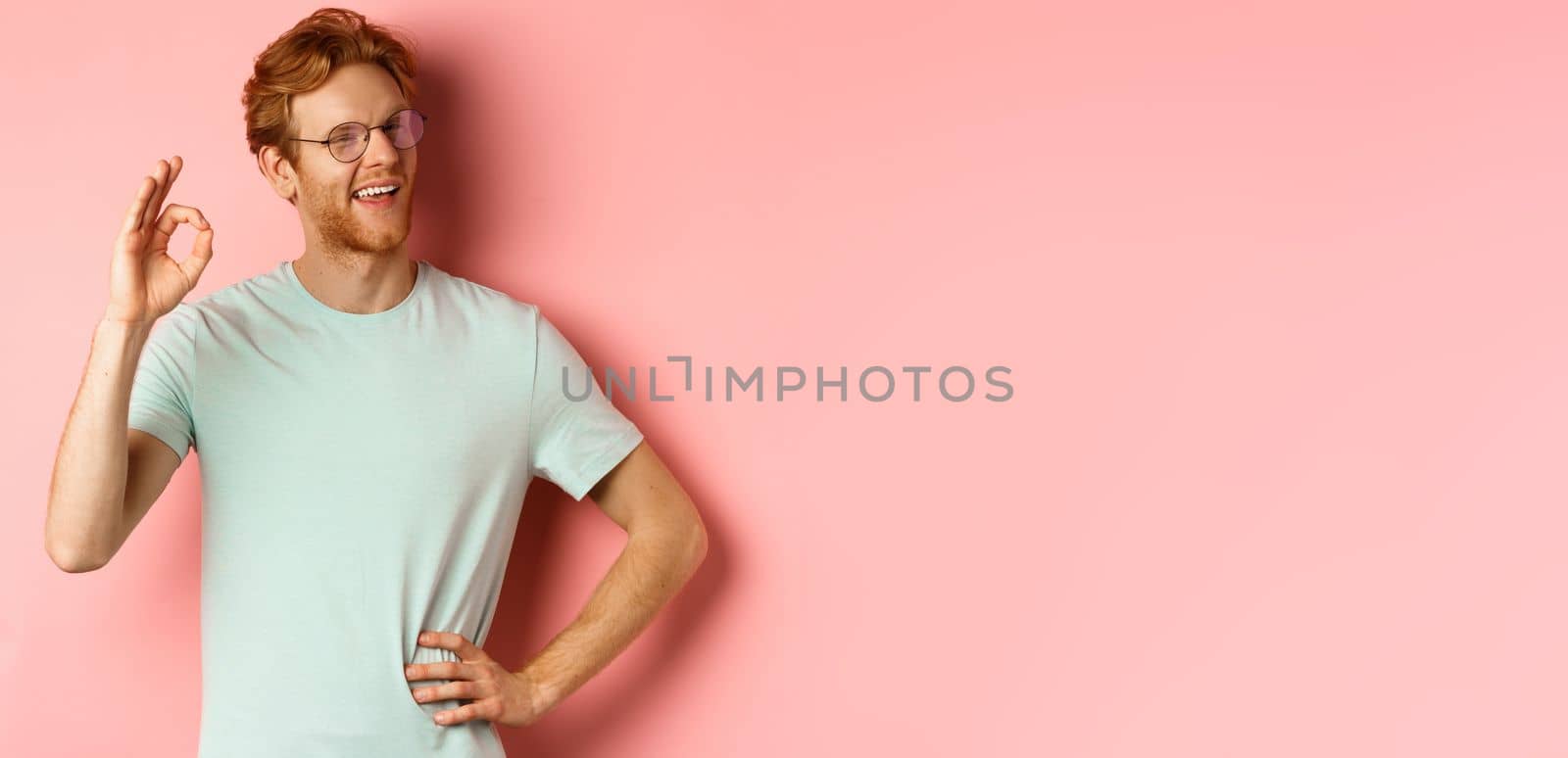 Cheerful guy with red hair and beard, wearing glasses, showing OK sign in approval and winking, smiling satisfied, standing over pink background by Benzoix
