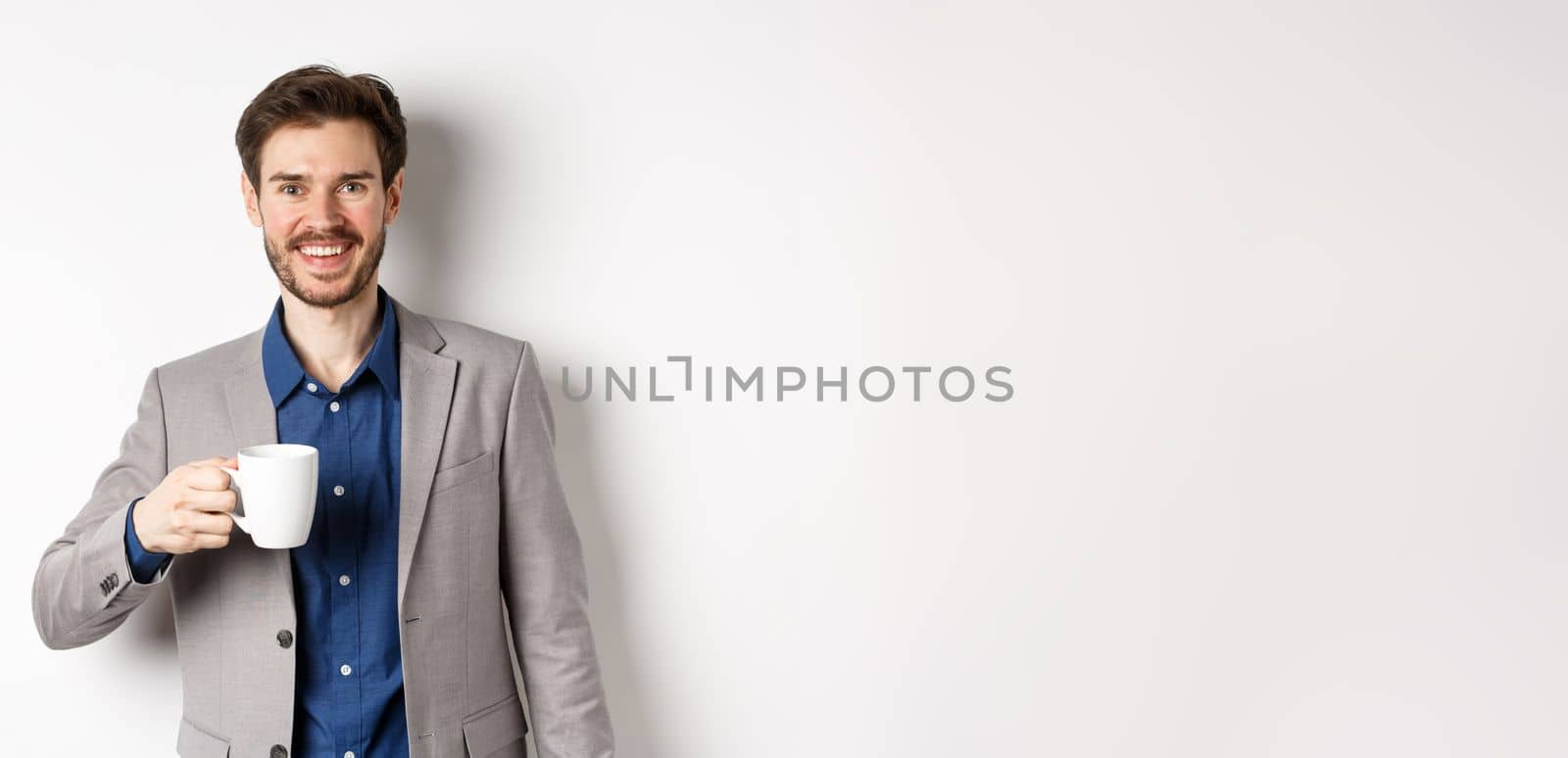 Handsome businessman in suit drinking coffee or tea from office mug, smiling enthusiastic at camera, standing against white background.